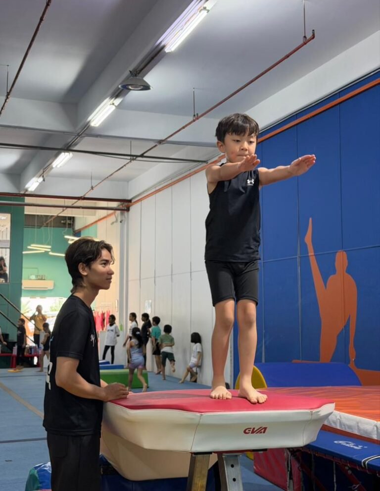 A coach guides a child on a balance beam at Flykidz Gymnastics in Petaling Jaya, showcasing kids gymnastics programs.