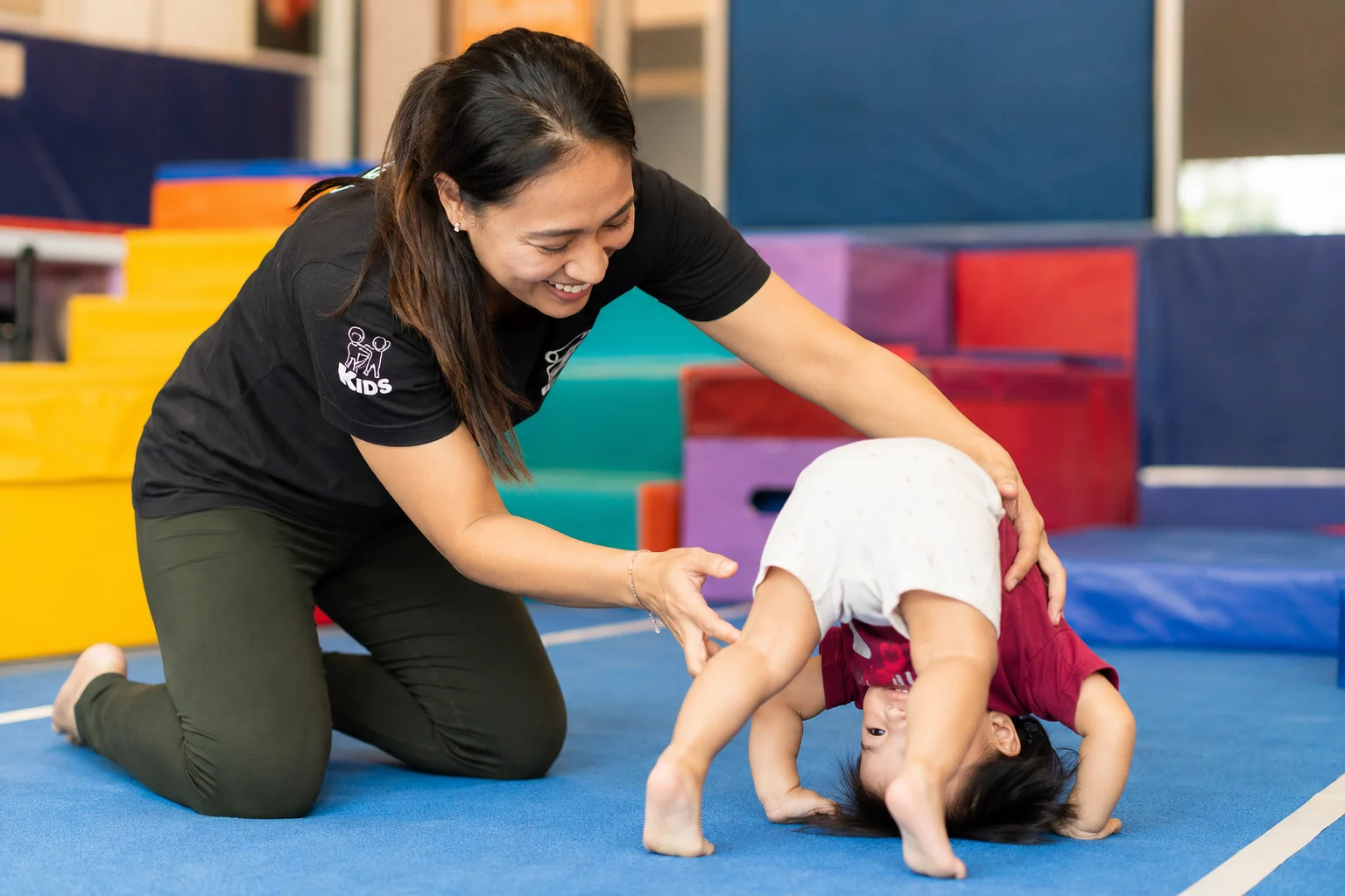 Flykidz coach guiding a toddler through a forward roll on the gymnastics mat, Petaling Jaya